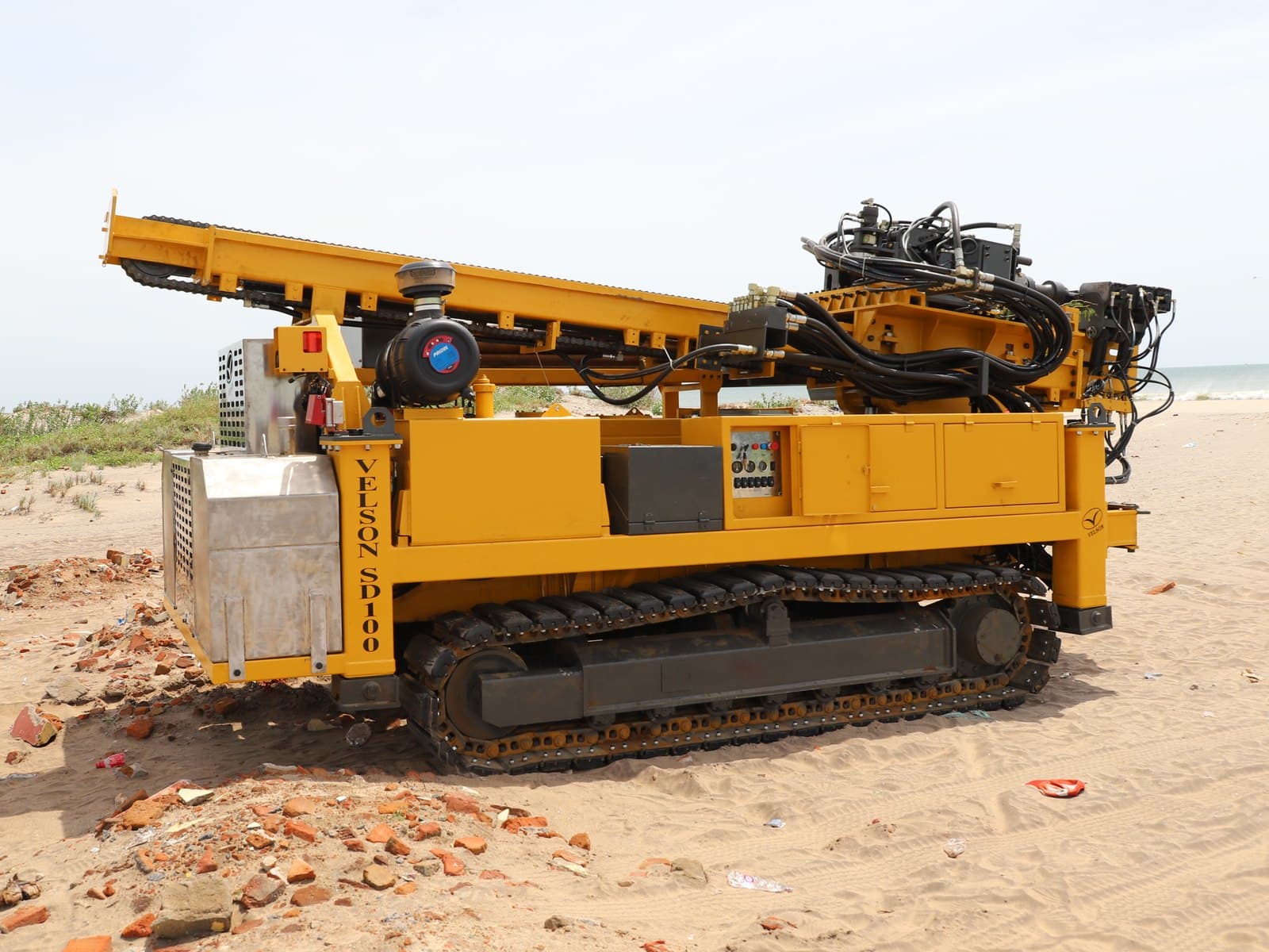 SAMP crawler sampler side-on on a coastal sand site, mast folded down along the deck, side control panel and hydraulic hose loom above the rotary head